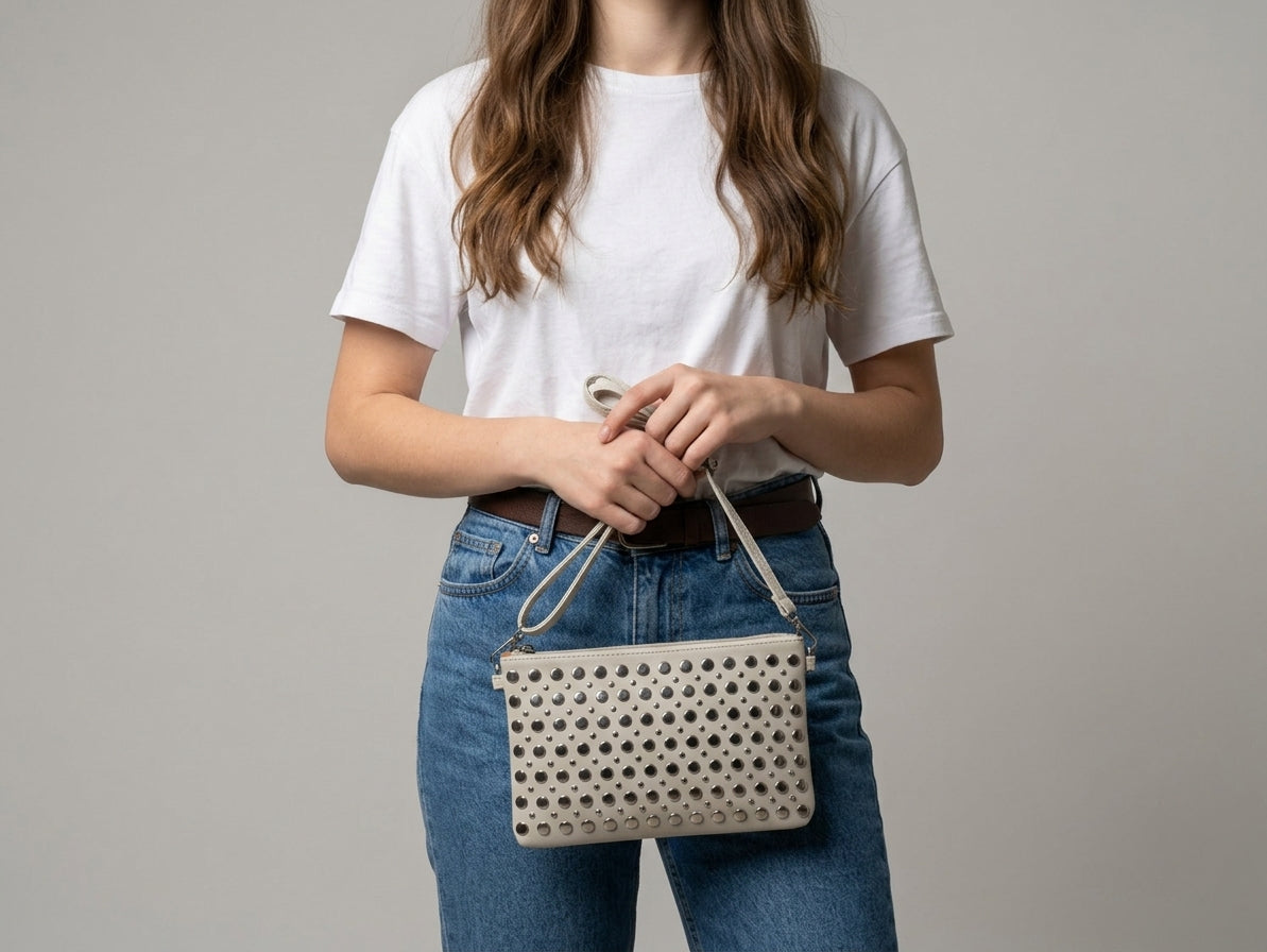 Mujer joven con camiseta blanca y jeans sujetando el Bolso Studded Cream 
de Sauga, bolso crema con tachuelas metálicas plateadas.